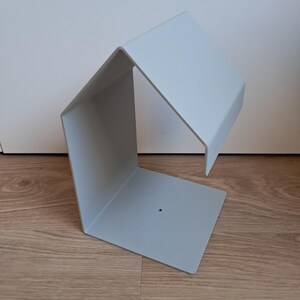 May include: A light gray, geometric metal shelf with a unique angular design. The shelf has a square base, a vertical side, and a horizontal top, creating an open, modern aesthetic. The shelf is made of metal and has a small hole on the base.