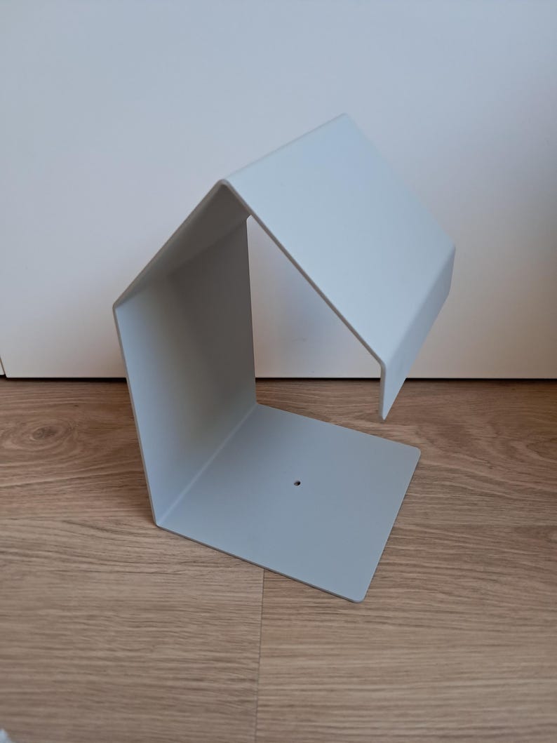 May include: A light gray, geometric metal shelf with a unique angular design. The shelf has a square base, a vertical side, and a horizontal top, creating an open, modern aesthetic. The shelf is made of metal and has a small hole on the base.