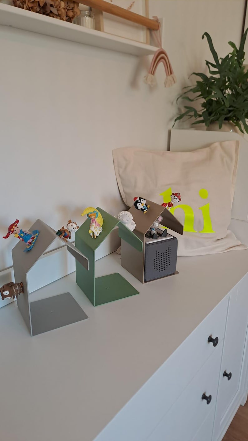 May include: Three metal house-shaped bookends in gray and green, each with a small figurine on the roof. A canvas pillow with the word "ki" in neon yellow is in the background.
