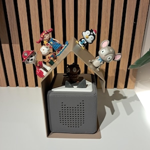 May include: A gray audio player with a speaker shaped like a cat's face, topped with a white lid. Several small figurines, including a penguin, a mouse, and a monkey, are arranged on a clear plastic roof structure above the player. The background features vertical wooden panels.