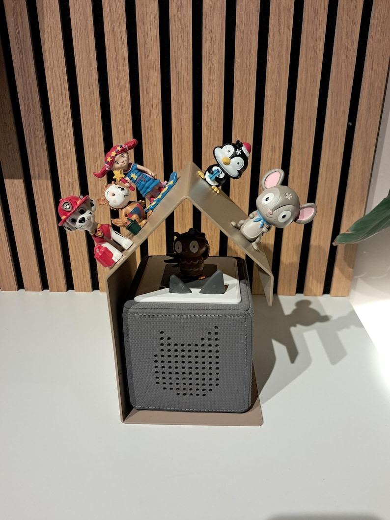 May include: A gray audio player with a speaker shaped like a cat's face, topped with a white lid. Several small figurines, including a penguin, a mouse, and a monkey, are arranged on a clear plastic roof structure above the player. The background features vertical wooden panels.