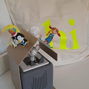 May include: A gray speaker with a house-shaped top, featuring small figurines of a penguin, mouse, and girl. A pillow with the word "hi" in neon yellow is in the background. The figurines are colorful and detailed.