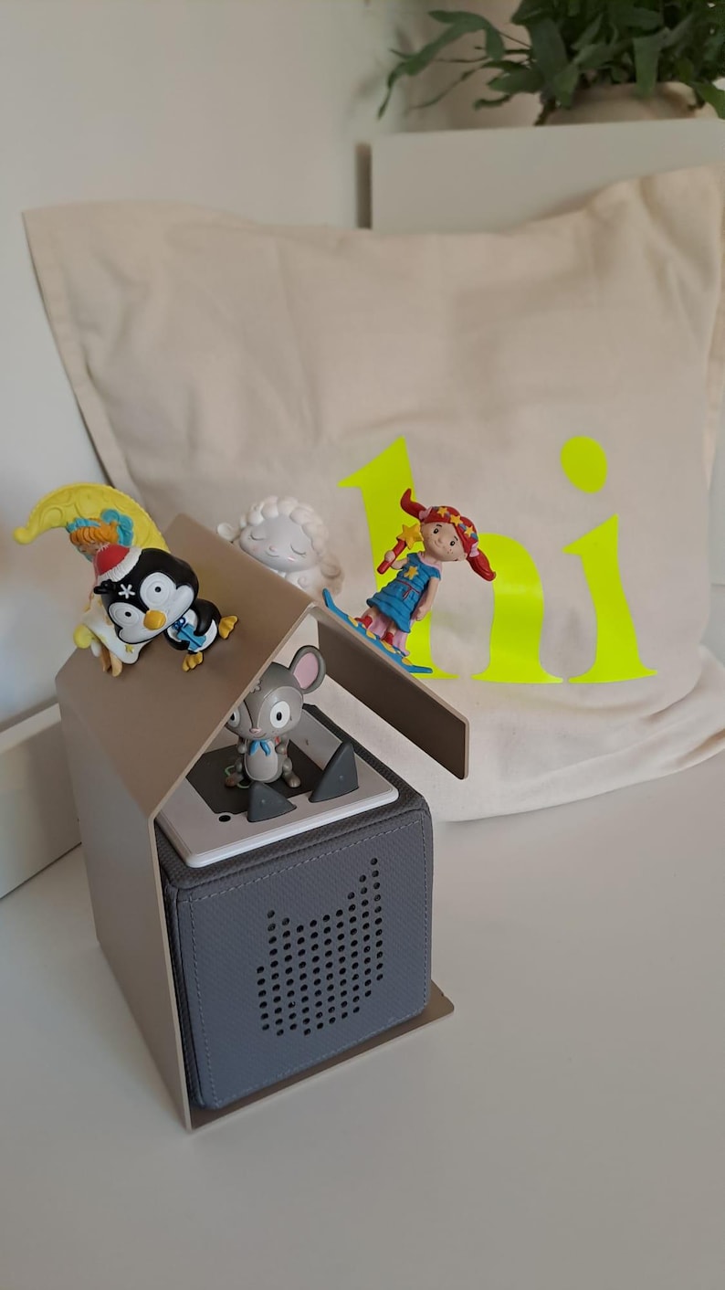 May include: A gray speaker with a house-shaped top, featuring small figurines of a penguin, mouse, and girl. A pillow with the word "hi" in neon yellow is in the background. The figurines are colorful and detailed.