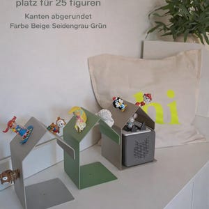 May include: A display shelf in the shape of a house, holding small figurines. The shelf is in shades of gray and green. The text on the wall says "Toniergal platz f&uuml;r 25 figuren".