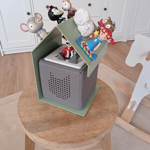 May include: A gray, cube-shaped speaker with a green roof and various toy figurines. The toys include a mouse, penguin, sheep, and a girl on a snowboard. The speaker sits on a wooden stool.