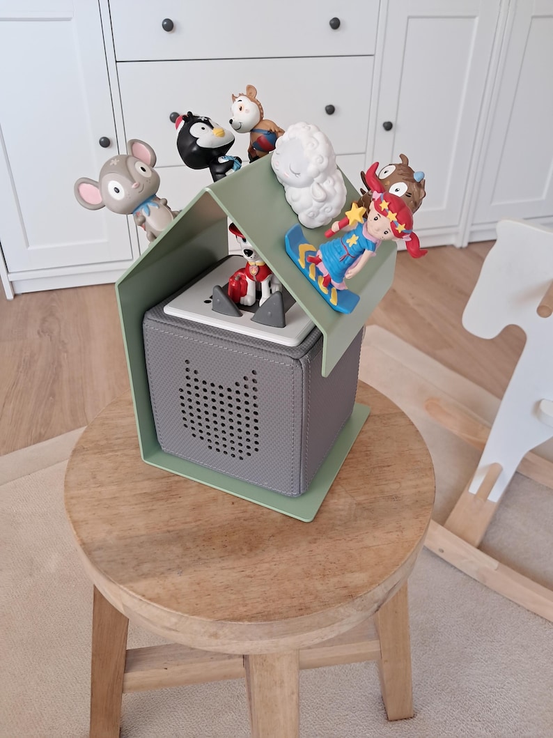 May include: A gray, cube-shaped speaker with a green roof and various toy figurines. The toys include a mouse, penguin, sheep, and a girl on a snowboard. The speaker sits on a wooden stool.