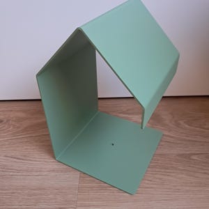 May include: A mint green metal bookend with a geometric design. The bookend has a rectangular base and a slanted top, creating an open, angular shape. The object is made of a single piece of metal and has a small hole on the base.