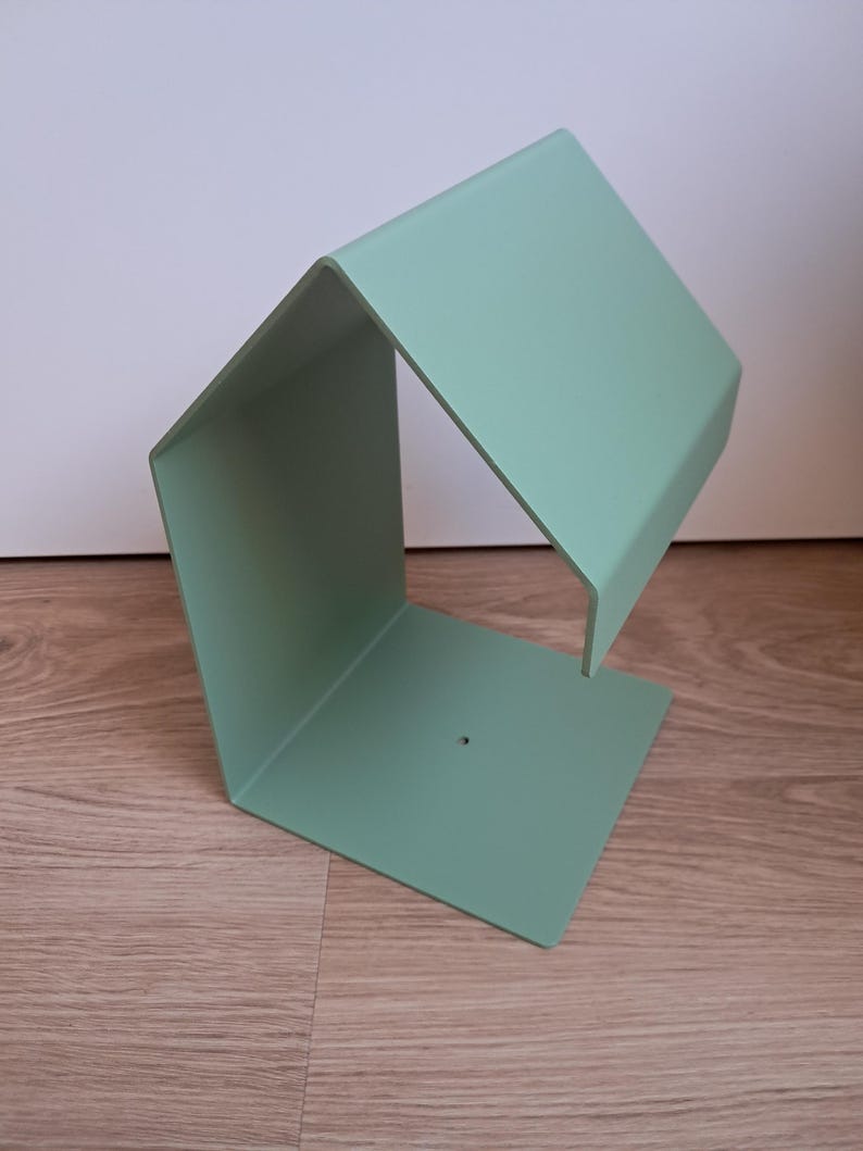 May include: A mint green metal bookend with a geometric design. The bookend has a rectangular base and a slanted top, creating an open, angular shape. The object is made of a single piece of metal and has a small hole on the base.
