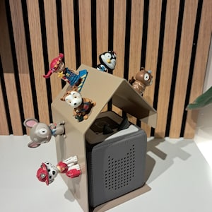 May include: A gray speaker with a perforated front panel is housed in a beige metal structure, resembling a house. Several small, colorful figurines of animals and characters are arranged around the structure.