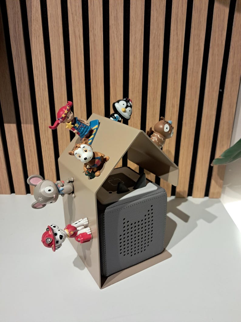 May include: A gray speaker with a perforated front panel is housed in a beige metal structure, resembling a house. Several small, colorful figurines of animals and characters are arranged around the structure.