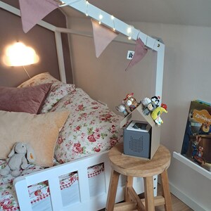 May include: A child's bedroom with a white bed frame, floral bedding, and decorative pillows. A small wooden stool holds a gray speaker with a cardboard house on top, decorated with small toys. Pink pennant flags and string lights adorn the bed frame.