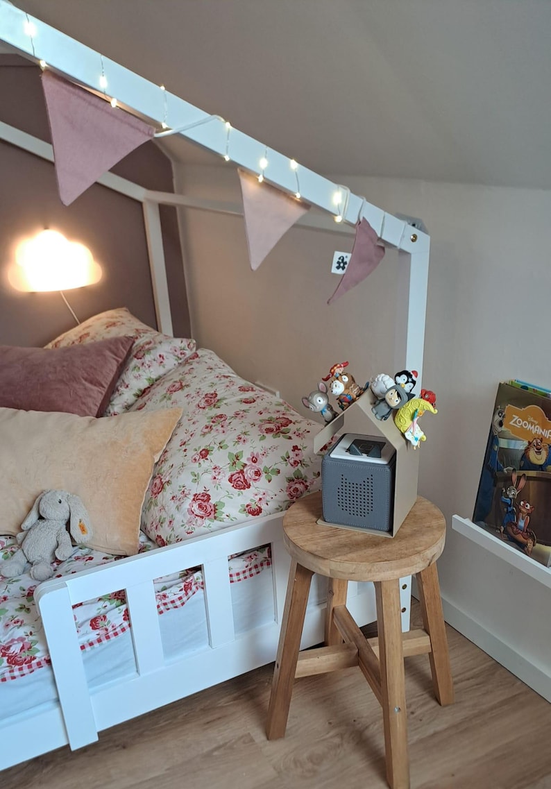 May include: A child's bedroom with a white bed frame, floral bedding, and decorative pillows. A small wooden stool holds a gray speaker with a cardboard house on top, decorated with small toys. Pink pennant flags and string lights adorn the bed frame.