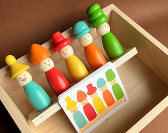 May include: Wooden toy set featuring six colorful peg dolls with expressive faces and matching hats, arranged in a wooden box. A card with matching illustrations is included, suggesting a matching game. The dolls are painted in various colors, including yellow, red, and green.