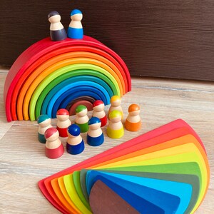 Pode incluir: Um brinquedo de empilhar de madeira arco-íris com 12 peças, incluindo um arco arco-íris e um conjunto de pessoas de madeira coloridas. O arco é feito de 10 peças de madeira, cada uma pintada de uma cor diferente do arco-íris. As pessoas são todas do mesmo tamanho e forma, mas cada uma tem um chapéu de uma cor diferente.