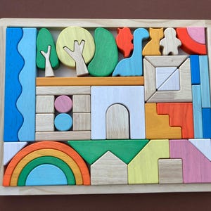 Può includere: Puzzle in legno con una varietà di forme colorate, tra cui un arcobaleno, alberi e un dinosauro. I pezzi del puzzle sono disposti all'interno di una cornice di legno chiaro, creando una scena vivace. Il puzzle è per bambini.