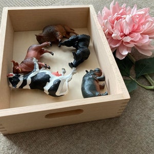 May include: A wooden box with a variety of animal figurines inside, including a cow, a bull, a hippopotamus, a rhino, and a black bear. The box is made of light wood and has a handle on the front.