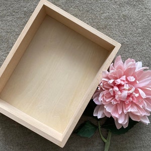 May include: A light wood rectangular box with a smooth surface. The box is empty and sits on a light brown textured surface. A pink flower with green leaves is partially visible in the lower right corner of the image.