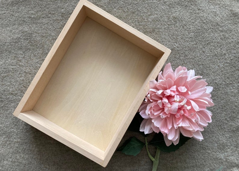 May include: A light wood rectangular box with a smooth surface. The box is empty and sits on a light brown textured surface. A pink flower with green leaves is partially visible in the lower right corner of the image.