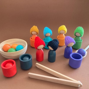Peut inclure: Ensemble de jouets en bois avec des poupées en bois colorées portant des bonnets tricotés, des bols et des pinces. L'ensemble comprend des boules orange, vertes et bleues, et des récipients rouges, bleu sarcelle et violets. Un jouet amusant et éducatif.
