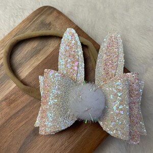 May include: A white and iridescent glitter bunny headband with a white pom-pom in the center. The headband is made of a brown elastic band.