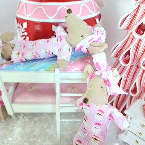 May include: A miniature Christmas scene featuring two mouse dolls in pink Santa Claus pajamas. One mouse is in a bunk bed, and the other is standing. A red and pink ornament and a candy cane tree are in the background.