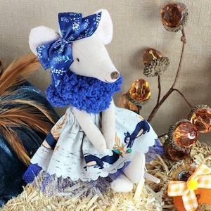 May include: A handmade fabric mouse doll with a blue bow, collar, and patterned dress. The dress features a deer design. The doll is sitting on straw with decorative fall-themed accents, including faux acorns and pumpkins.