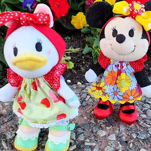 May include: Two plush dolls of cartoon characters, Daisy Duck and Minnie Mouse, stand side-by-side. Daisy Duck wears a red bow, a green dress with strawberries, and mint green shoes. Minnie Mouse wears a black bow, a floral dress, and red shoes.