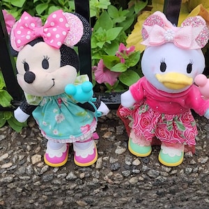 May include: Two plush character dolls, Minnie Mouse and Daisy Duck, stand side-by-side. Minnie wears a pink polka-dot bow, a turquoise floral dress, and pink shoes. Daisy dons a pink rose-patterned dress and matching shoes. The dolls are set against a backdrop of greenery.