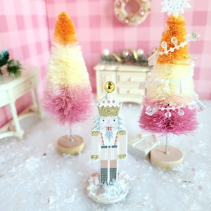 May include: A festive holiday scene featuring a wooden nutcracker figurine and two ombre bottle brush Christmas trees. The trees transition from pink to yellow to orange. A snowflake and beads adorn one tree. The nutcracker is white with gold accents.