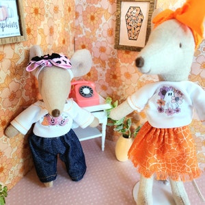 May include: Two fabric mice in Halloween outfits. One wears a white top with a pumpkin design and blue jeans. The other wears a white top with a ghost design and an orange skirt. The background has floral wallpaper.