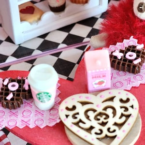 May include: Miniature bakery scene with a white display case filled with tiny pastries, including tarts and cupcakes. A red table holds a coffee cup, chocolate waffles with pink accents, and a wooden heart-shaped coaster. A pink milk carton is also present.