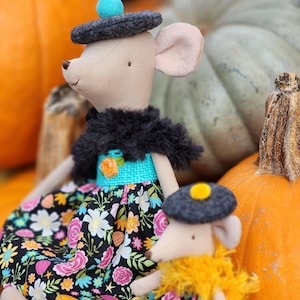 May include: Two plush mice dolls, one large and one small, are dressed in floral print dresses and gray hats. The larger mouse has a black fur collar and a turquoise waistband. The background features orange and green pumpkins.