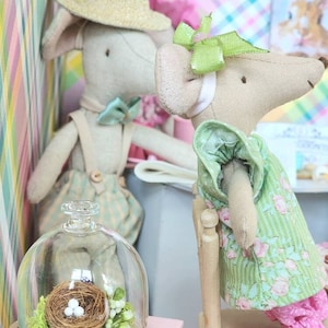 May include: Two fabric mouse dolls, one wearing a straw hat and bow tie, the other in a green floral dress and pink pants. A glass cloche displays a miniature bird's nest with eggs and greenery. The scene evokes a whimsical, handcrafted aesthetic.