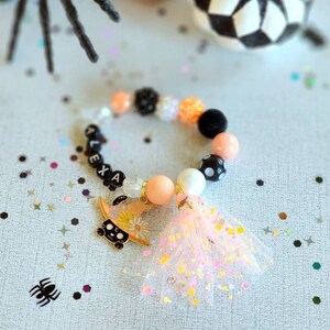 Puede incluir: Una pulsera de cuentas con el nombre "ALEXA" escrito con cuentas de letras negras. La pulsera presenta un dije de un gato negro con un sombrero de bruja y una borla rosa. Las cuentas son en tonos melocotón, blanco, negro y naranja.