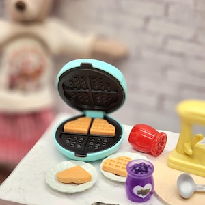May include: Miniature toy waffle maker with waffles, plates, and a purple jar with a heart design. The waffle maker is light blue and black. The waffles are golden brown. A red shaker and a spoon are also present.