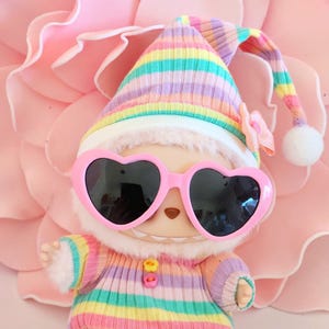 May include: A whimsical doll wearing pink heart-shaped sunglasses, a striped knit hat, and a matching striped outfit in pastel colors. The doll has a white furry trim and a small bow. The background is a large pink flower.