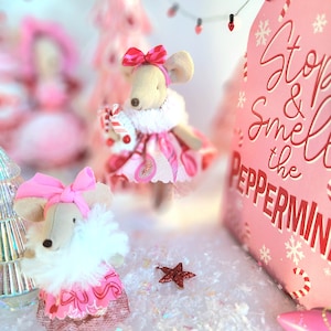 May include: A whimsical holiday scene featuring plush mice dressed in pink dresses and bows, with white fur collars. A pink sign reads "Stop & Smell the Peppermint." Decorative Christmas trees and a red star add to the festive atmosphere.