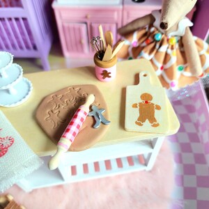May include: A miniature kitchen scene with a light-colored table, rolling pin, and gingerbread man-shaped cutting board. A gingerbread man is on the cutting board. A doll is in the background. The scene is decorated in pink and white.