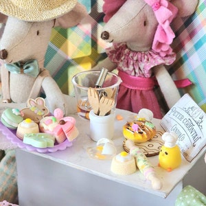 May include: A whimsical scene featuring two mouse dolls, one wearing a straw hat and the other a pink headpiece. Miniature pastries, a rolling pin, and a chef chick figurine are arranged on a white surface, creating a charming bakery setting.