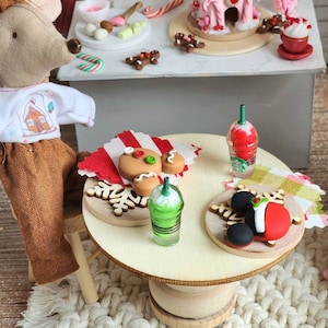 May include: A miniature Christmas scene featuring a mouse doll and a table with festive treats. The table holds gingerbread cookies, a green drink, and a red and white dessert. A gingerbread house and other sweets are on a higher surface.