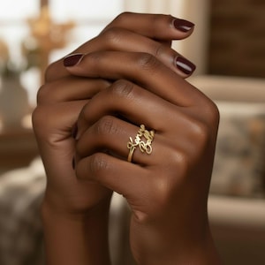 May include: A gold ring inscribed with the name "Kenny Belle" in a cursive font. The ring is worn on a finger, with the hands clasped together. The nails are painted a dark burgundy colour.