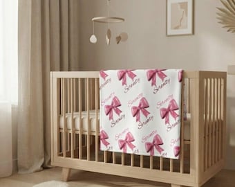May include: A white baby blanket featuring a repeating pattern of glossy pink bows and the name Serenity written in a cursive script. The blanket is draped over the side of a light-colored wooden crib in a room with neutral-toned decor.