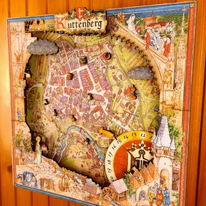May include: A detailed, colorful 3D map of Kuttenberg, featuring a clock and illustrations of buildings and people. The map is framed and mounted on a wooden wall, showcasing a historical town layout with intricate details.