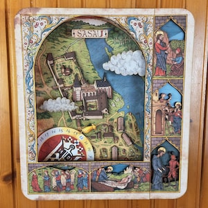 May include: A rectangular wooden wall art piece featuring a multi-layered 3D map of Sasau with a central castle, a blue river, and white clouds. The border displays intricate floral patterns and three vertical panels depicting religious figures.
