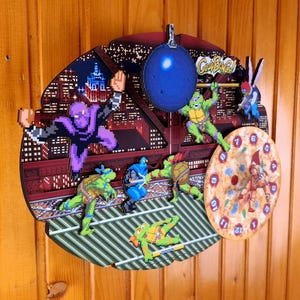 May include: A decorative wall clock featuring a cityscape backdrop with pixelated characters in action poses. The clock includes a pizza-themed clock face and the words "Cowabunga!" in a banner.