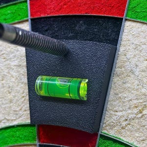 May include: A close-up shot of a dartboard with a dart embedded in the black surface. A small, bright green level is attached to the board. The dartboard has red, black, green, and beige sections.