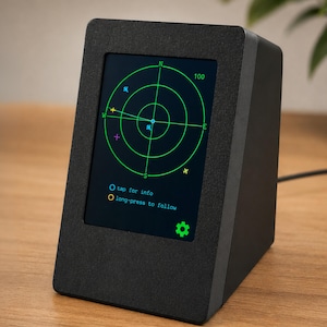 Puede incluir: Un dispositivo electrónico negro en forma de cuña con una pantalla digital que muestra una interfaz de radar verde. La pantalla muestra puntos cardinales, iconos de aviones y el texto: tap for info, long-press to follow, y un icono de engranaje.