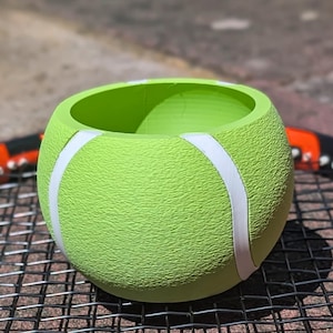 Peut inclure: Un pot de fleurs vert citron en forme de balle de tennis avec des rayures blanches. Le pot a une surface texturée et une ouverture ronde. Le pot est posé sur une raquette de tennis.