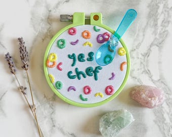 May include: A green embroidery hoop with the words "yes chef" stitched in teal thread. The design includes colorful embroidered shapes. A blue spoon rests on the hoop. Lavender sprigs and crystals are also present.