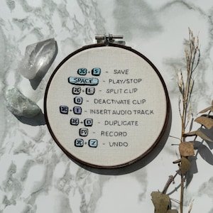 May include: A white embroidery hoop with brown wood framing. The hoop features a list of keyboard shortcuts for audio editing software, including "Save", "Play/Stop", "Split Clip", "Deactivate Clip", "Insert Audio Track", "Duplicate", "Record", and "Undo".